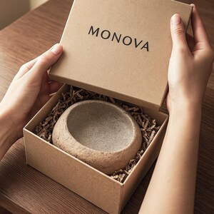 May include: A small, round, stone-like bowl in a cardboard box with the word "MONOVA" on the lid. The box contains brown paper shreds. The bowl has a rough, textured surface. The box is being opened.