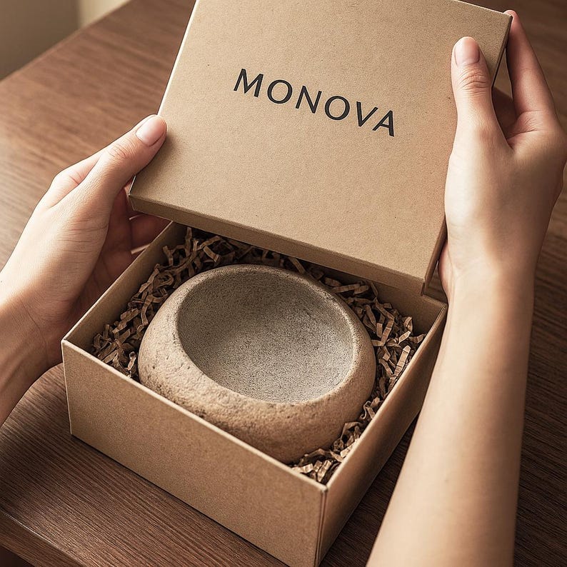 May include: A small, round, stone-like bowl in a cardboard box with the word "MONOVA" on the lid. The box contains brown paper shreds. The bowl has a rough, textured surface. The box is being opened.