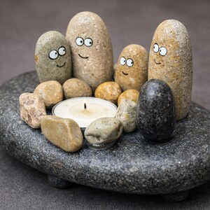 May include: A unique candle holder made of natural stones. The centerpiece features a lit candle surrounded by smaller stones. Several larger, oblong stones with painted faces create a whimsical, friendly design. The base is a large, gray stone.