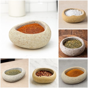 Może przedstawiać: Kolekcja małych, kamiennych misek wypełnionych różnymi przyprawami. Przyprawy obejmują płatki czerwonej papryki chili, zielone zioła i pomarańczową kurkumę. Miski mają naturalny, organiczny kształt i fakturę.