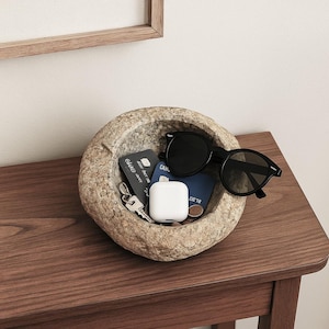 May include: A textured stone bowl holds various items on a wooden table. Inside are black sunglasses, keys, a white earbud case, and cards. The bowl's surface is rough, contrasting with the smooth contents. The table is made of dark wood.
