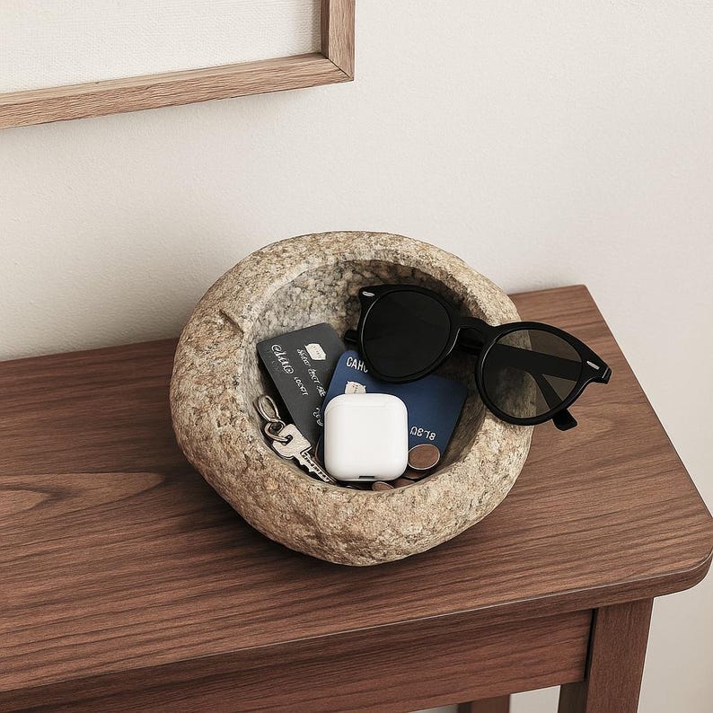 May include: A textured stone bowl holds various items on a wooden table. Inside are black sunglasses, keys, a white earbud case, and cards. The bowl's surface is rough, contrasting with the smooth contents. The table is made of dark wood.