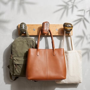 May include: A light oak wall-mounted rack featuring three distinct stone-shaped hooks. Hanging from the hooks are an olive green canvas backpack, a smooth tan leather tote bag with dual handles, and a plain off-white cotton canvas tote bag.