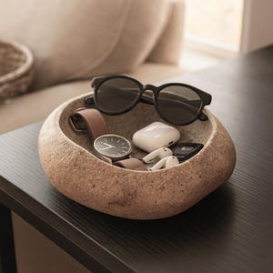 May include: A beige stone bowl with a textured surface contains accessories. Black sunglasses sit on top. Inside are a wristwatch with a brown leather strap, white wireless earbuds, and a black car key fob. The bowl is on a dark surface.