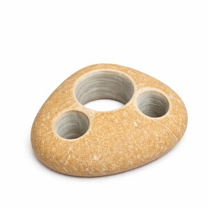 May include: A natural stone toothbrush holder with three circular openings. The holder is a light tan color with a textured surface and a smooth interior in the openings. The design is organic and minimalist.