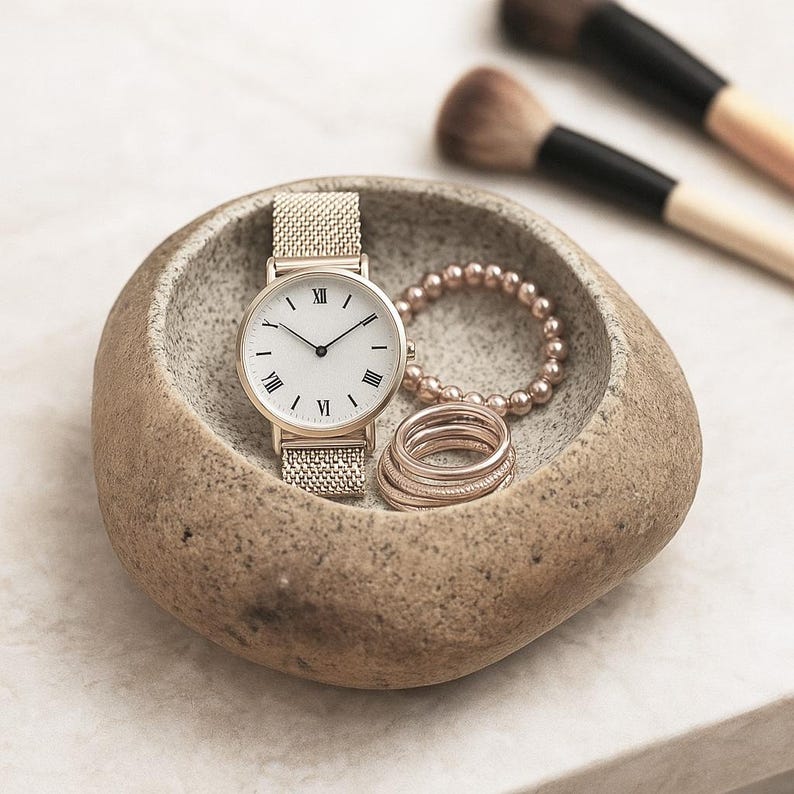 May include: A gold wristwatch with a mesh strap and a white face with Roman numerals rests in a stone bowl. A beaded bracelet and rings are also in the bowl. Makeup brushes are in the background.
