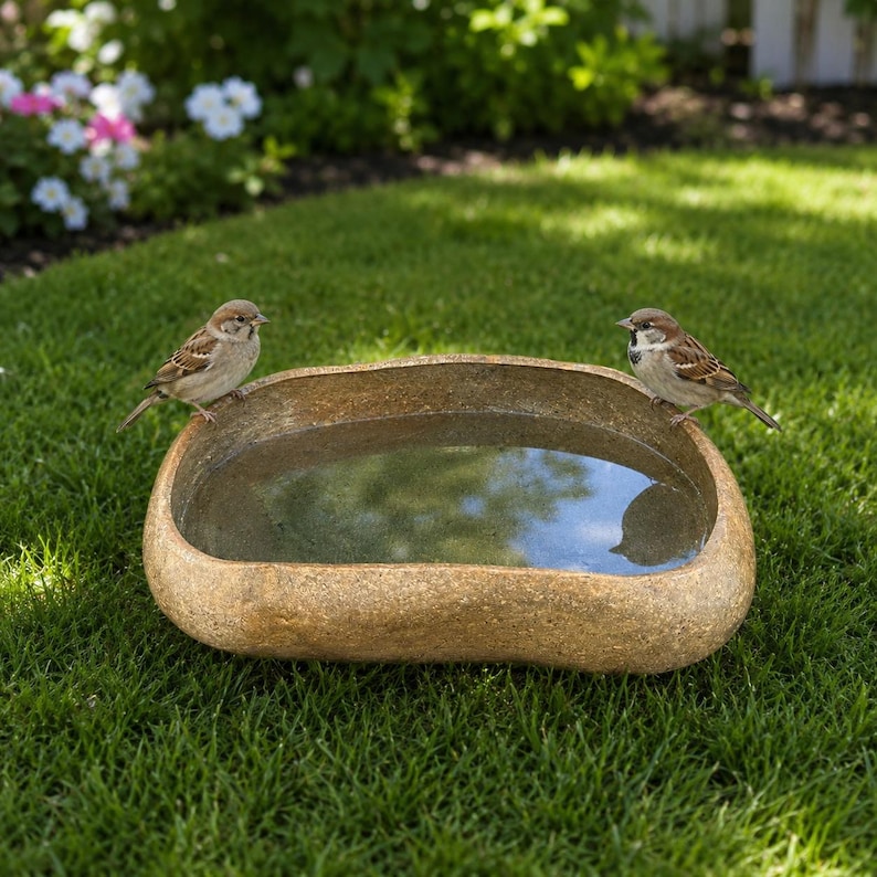 May include: A low, tan, stone-textured bird bath basin filled with clear water. Two small brown and grey sparrows are perched on opposite sides of the rim, facing inward toward the center of the basin, which reflects the surrounding environment.