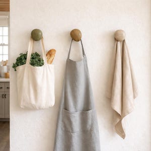 Può includere: Tre ganci a muro con oggetti appesi. Una borsa di tela piena di cavolo riccio e una baguette, un grembiule grigio e un asciugamano beige. I ganci sono rotondi e di pietra.