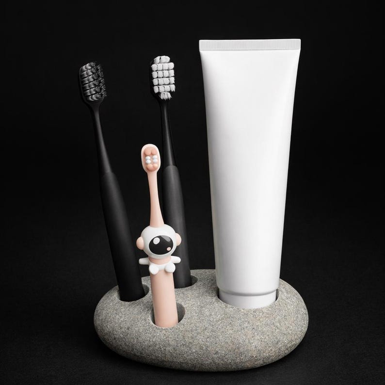 May include: A gray stone toothbrush holder with three black toothbrushes, a pink astronaut-themed toothbrush, and a white toothpaste tube. The toothbrushes have different colored bristles. The holder is on a black background.