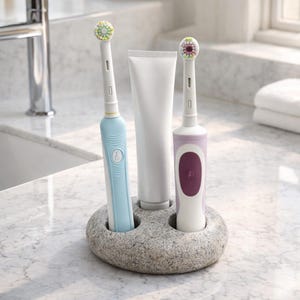 Op de afbeelding: Een lichtblauwe en een lavendelkleurige elektrische tandenborstel met een tube tandpasta in een grijze stenen houder. De tandenborstels hebben witte borstelkoppen met kleurrijke borstelharen. De houder staat op een marmeren aanrecht.