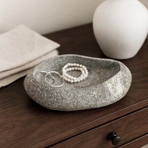 May include: A grey stone-effect jewellery dish contains a pearl bracelet, silver hoop earrings, and a delicate necklace. The dish is on a dark wood surface, with a white vase in the background. A folded linen cloth is also visible.