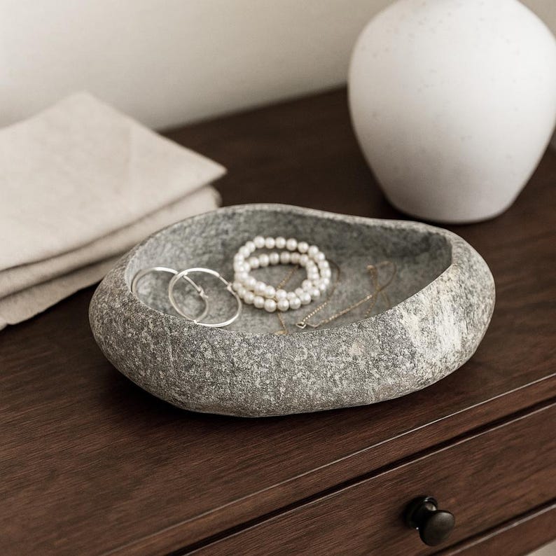 May include: A grey stone-effect jewellery dish contains a pearl bracelet, silver hoop earrings, and a delicate necklace. The dish is on a dark wood surface, with a white vase in the background. A folded linen cloth is also visible.