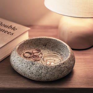 May include: A grey stone jewellery dish containing gold rings and bracelets, including a ring with two small pearls. The dish is on a wooden surface, near a book and a lamp with a beige lampshade.