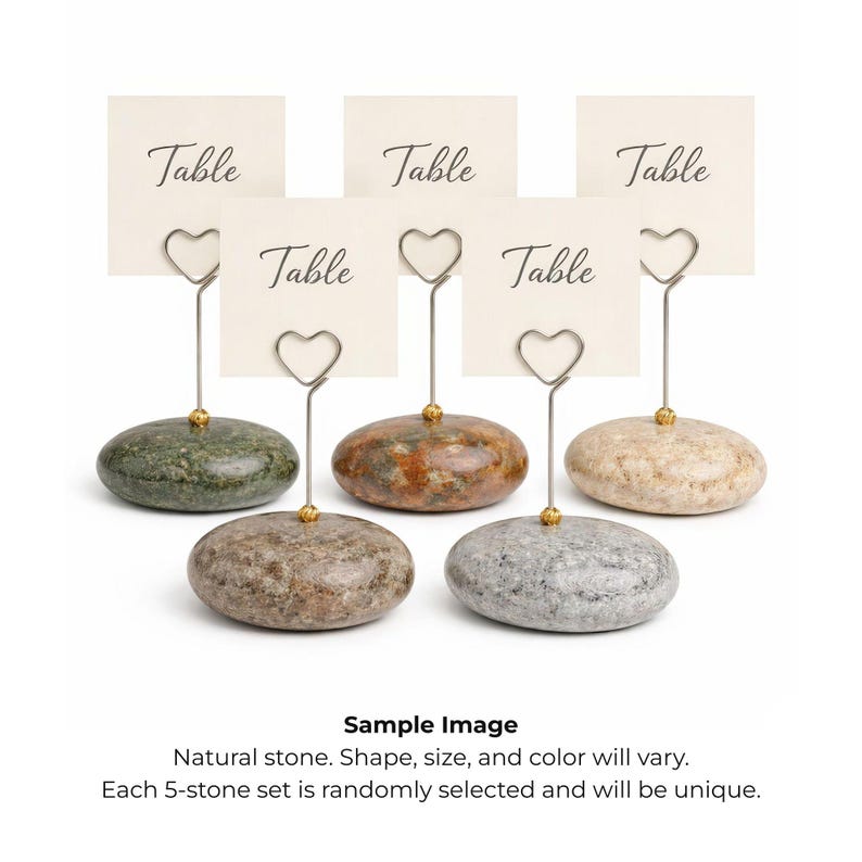 May include: A set of five natural stone table card holders. Each holder features a unique stone base with a silver-colored wire that forms a heart shape to hold a card. The cards read "Table".