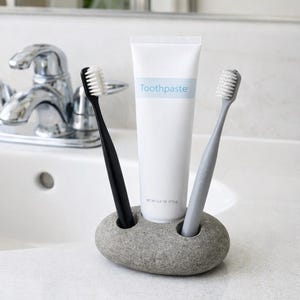 Può includere: Un portaspazzolini in pietra grigia con uno spazzolino nero e uno grigio, e un tubetto di dentifricio. Il tubetto di dentifricio è bianco con la scritta "Toothpaste" in blu. Il supporto è su un piano di lavoro bianco.