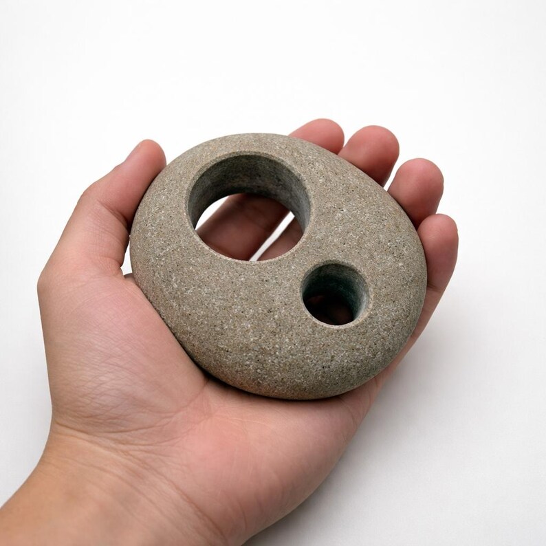 May include: A gray, smooth stone with two circular holes. The larger hole is near the top, and a smaller hole is below it. The stone is held in a hand against a white background. The stone's shape is oval.