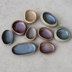 May include: A collection of nine small, smooth, pebble-shaped ceramic bowls in various earthy tones including brown, gray, tan, and dark blue. Each bowl features a rounded, organic form with a concave center, arranged in a scattered cluster.