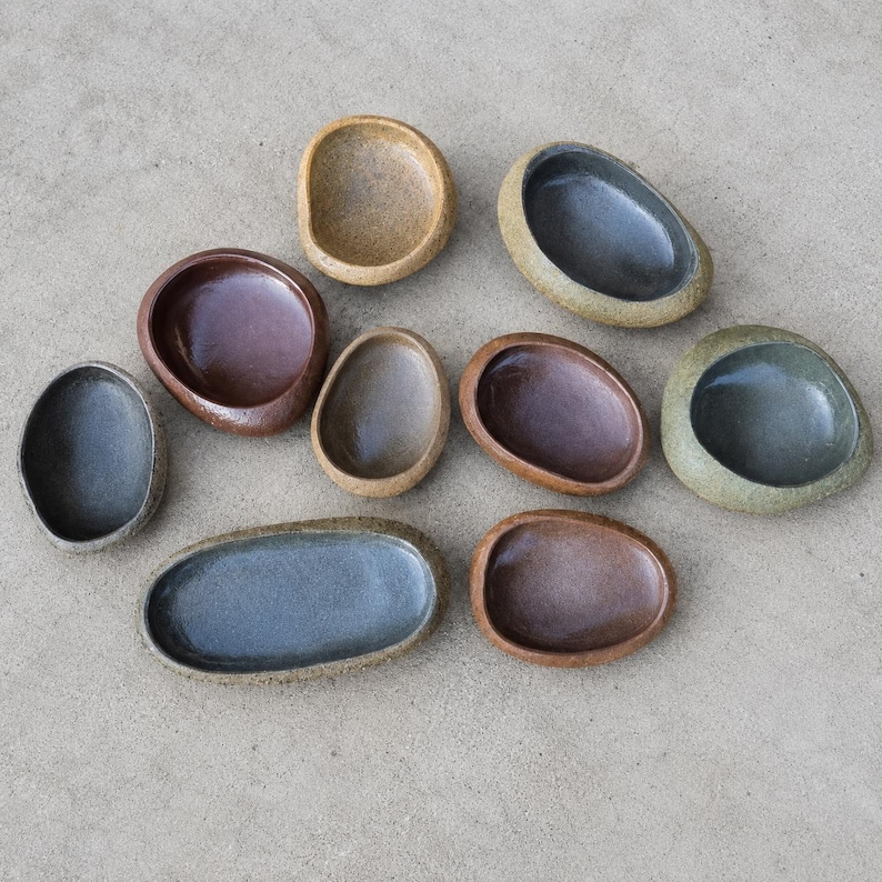 May include: A collection of nine small, smooth, pebble-shaped ceramic bowls in various earthy tones including brown, gray, tan, and dark blue. Each bowl features a rounded, organic form with a concave center, arranged in a scattered cluster.