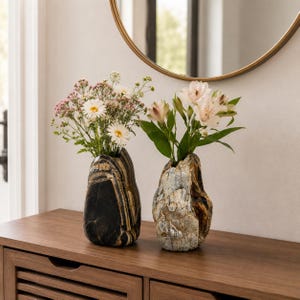 Peut inclure: Deux vases en pierre contenant des fleurs. Le vase de gauche est gris foncé avec des bandes horizontales brun clair. Le vase de droite est texturé, gris clair et brun avec un aspect vieilli. Les deux ont des formes organiques irrégulières.