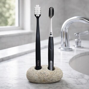 May include: Two toothbrushes, one black and one black and silver, are displayed in a stone holder. The black toothbrush has white bristles and the other has black bristles. The holder is a light tan color and sits on a marble surface.