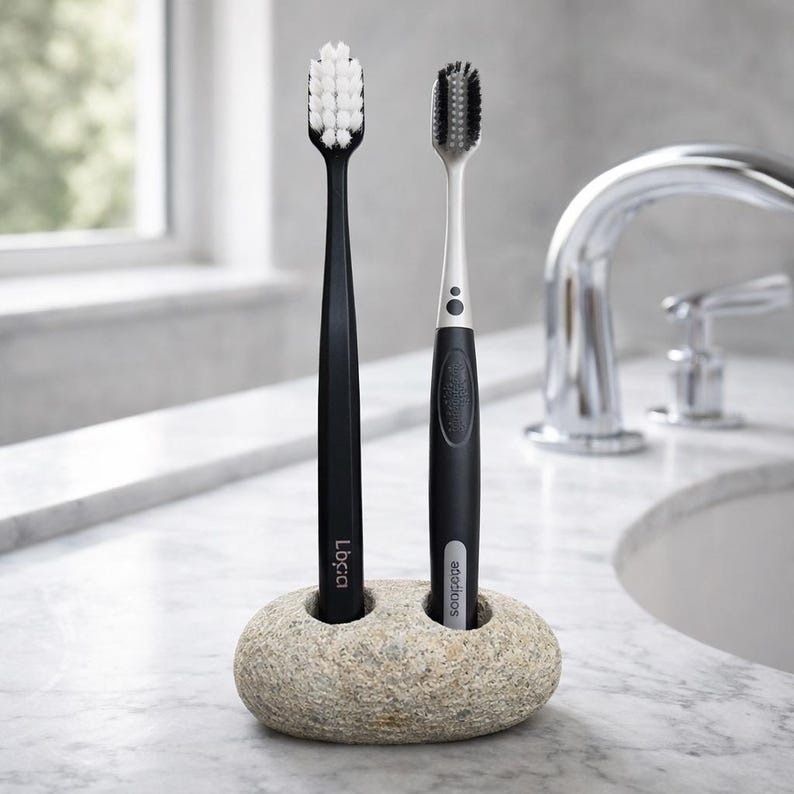 May include: Two toothbrushes, one black and one black and silver, are displayed in a stone holder. The black toothbrush has white bristles and the other has black bristles. The holder is a light tan color and sits on a marble surface.