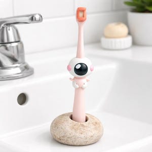 May include: A pink toothbrush with an orange head and a cartoon eye-shaped design. The toothbrush is in a beige stone holder. The design includes a large black eye with pink accents and small arms. The toothbrush is on a white sink.