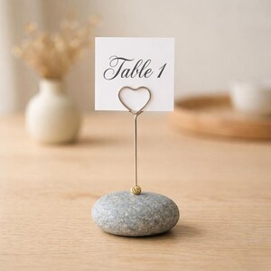 May include: A gray stone table number holder with a heart-shaped wire and a small gold bead. The white card reads "Table 1" in elegant script. The holder is on a light wood surface.