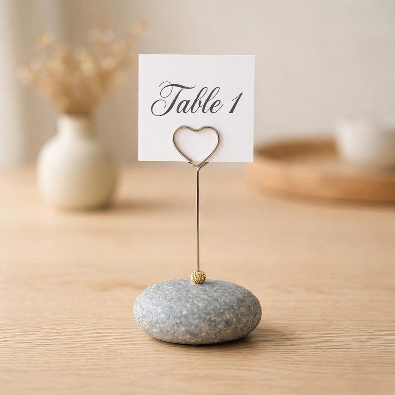 May include: A gray stone table number holder with a heart-shaped wire and a small gold bead. The white card reads "Table 1" in elegant script. The holder is on a light wood surface.
