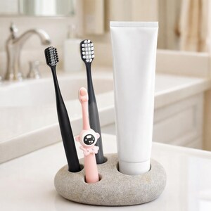 May include: A gray stone toothbrush holder with three toothbrushes and a tube of toothpaste. Two black toothbrushes and a pink toothbrush with a cartoon face are in the holder. The white toothpaste tube is upright.