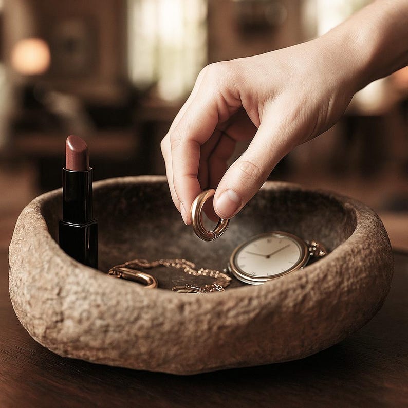 May include: A stone-effect bowl contains a gold earring, lipstick, a pocket watch, and a gold chain. A hand is reaching for the earring. The items are on a dark wooden surface, with a blurred background.