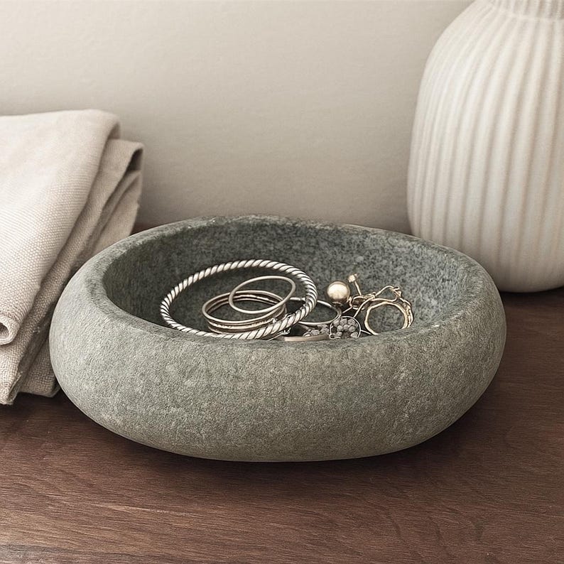 May include: A grey stone bowl containing jewellery, including silver bracelets and rings, and a pearl ring. The bowl is on a wooden surface, with a folded beige cloth and a white vase in the background. The bowl has a textured surface.