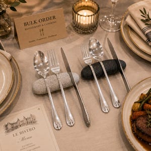 Könnte beinhalten: Tischarrangement mit Silberbesteck, Tellern und einer brennenden Kerze. Das Besteck umfasst Löffel, Gabeln und Messer. Eine Karte trägt die Aufschrift "BULK ORDER RESTAURANT & CATERING". Eine Speisekarte von "LE BISTRO" ist ebenfalls sichtbar.
