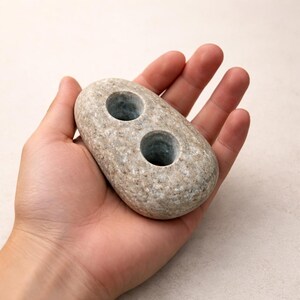 May include: A gray stone candle holder with two circular openings. The oblong-shaped holder is held in a hand, showcasing its natural texture and design. The candle holder is a decorative item.