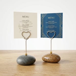Op de afbeelding: Twee menukaarthouders met hartvormige draadclips. Eén kaart is crèmekleurig met menu-items, de andere is marineblauw met gouden geometrische ontwerpen. Beide kaarten worden vastgehouden door een draad die aan een stenen voet is bevestigd.
