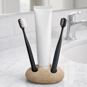 May include: A beige stone toothbrush holder with two black toothbrushes and a white tube of toothpaste. The toothbrushes have white and black bristles. The holder is on a marble countertop.