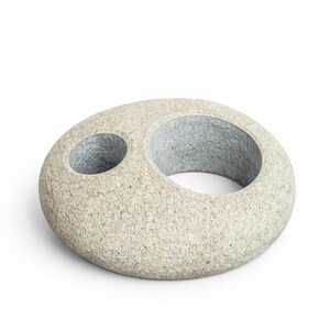 May include: A gray stone sculpture with a large circular opening and a smaller, adjacent hole. The stone has a speckled texture and a smooth, rounded shape. The sculpture is likely a decorative item for a home or garden.