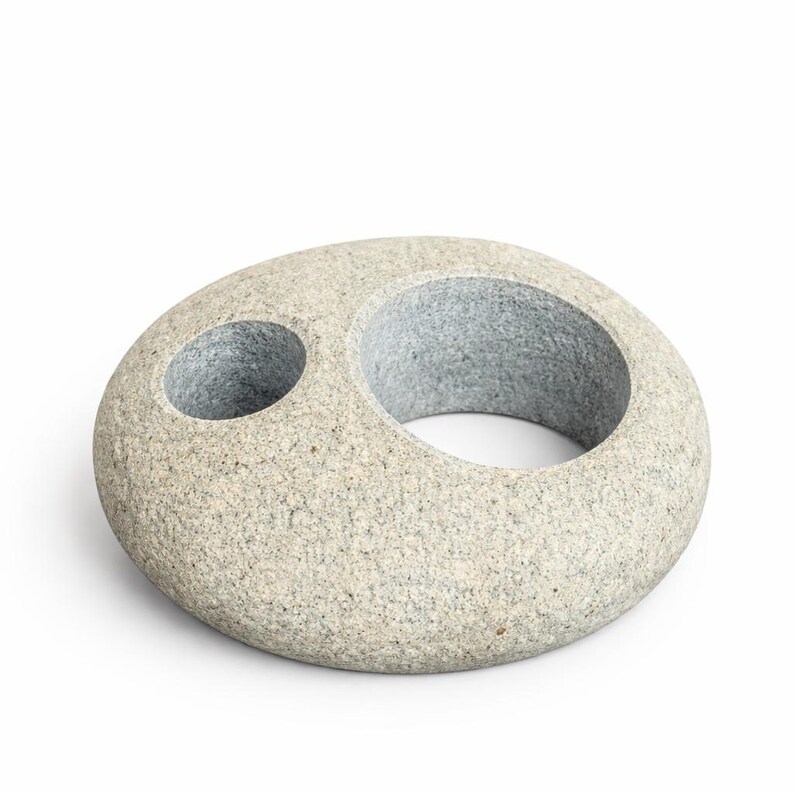 May include: A gray stone sculpture with a large circular opening and a smaller, adjacent hole. The stone has a speckled texture and a smooth, rounded shape. The sculpture is likely a decorative item for a home or garden.