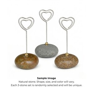 May include: Three natural stone card holders. Each holder features a silver-colored wire heart design attached to a stone base. The stones vary in color, including brown and gray, and have a small gold-colored accent.