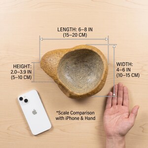May include: A beige, organic-shaped bowl with a textured surface. The bowl's length is 6-8 inches (15-20 cm), width is 4-6 inches (10-15 cm), and height is 2.0-3.9 inches (5-10 cm). A white iPhone is next to the bowl for scale comparison. The text reads "Scale Comparison with iPhone & Hand."