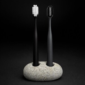 May include: Two black toothbrushes with white and black bristles are displayed in a gray stone holder. The toothbrushes stand upright against a black background. The holder has two circular openings.