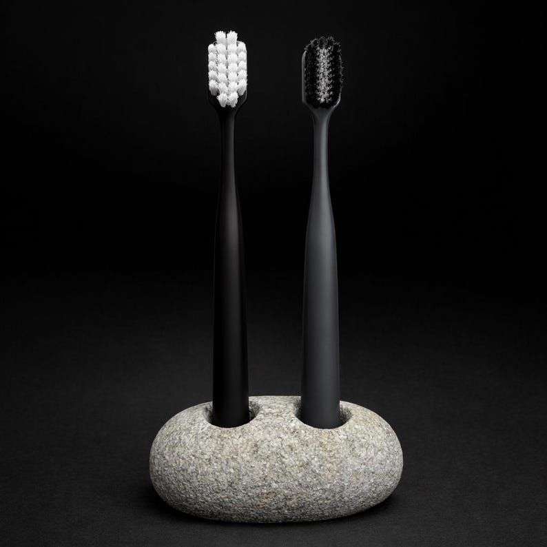 May include: Two black toothbrushes with white and black bristles are displayed in a gray stone holder. The toothbrushes stand upright against a black background. The holder has two circular openings.