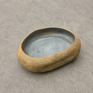 May include: An oval-shaped bowl with a textured, tan, stone-like exterior and a smooth, dark grey interior. The vessel features thick, rounded edges and a deep, open basin, presenting a natural, organic form with a contrasting color palette.