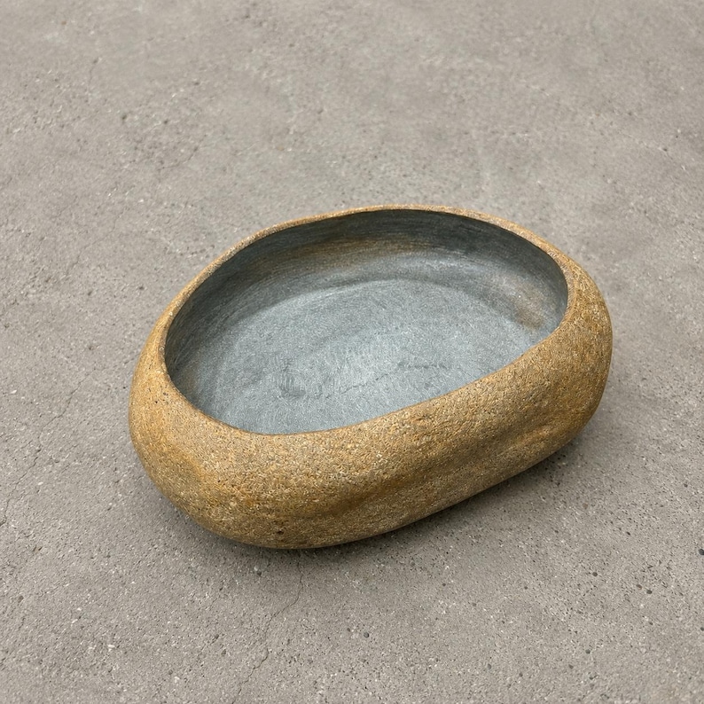 May include: An oval-shaped bowl with a textured, tan, stone-like exterior and a smooth, dark grey interior. The vessel features thick, rounded edges and a deep, open basin, presenting a natural, organic form with a contrasting color palette.