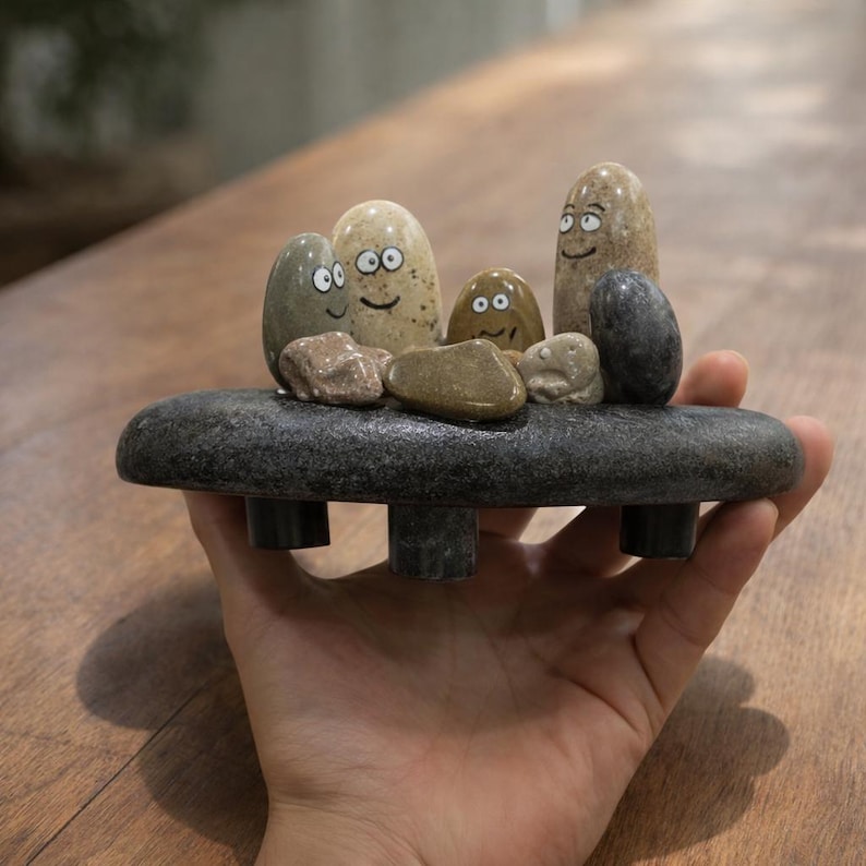 May include: A whimsical stone sculpture featuring a family of smiling rock figures. The figures are various sizes and colors, resting on a dark gray stone base with three short legs. The sculpture is held in a hand.