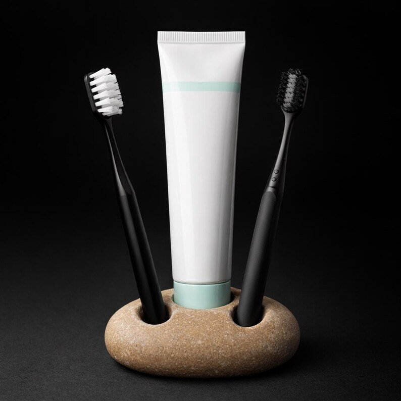 May include: A dental hygiene set featuring a white tube of toothpaste with a light blue stripe, two black toothbrushes, and a beige stone holder. One toothbrush has white bristles, the other has black bristles. The background is black.