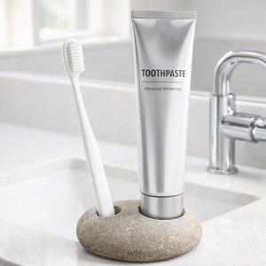 Puede incluir: Un cepillo de dientes blanco y un tubo de pasta dental con la etiqueta "TOOTHPASTE Advanced Whitening" se muestran en un soporte de piedra gris. Los art&iacute;culos est&aacute;n sobre una encimera blanca cerca de un lavabo y un grifo.