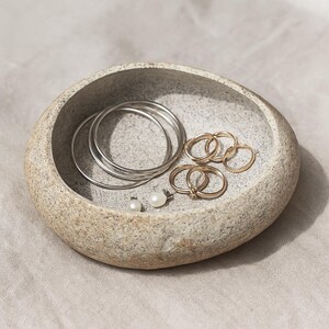 May include: A grey stone-effect jewellery dish contains silver bangles, gold rings, and pearl earrings. The dish has a textured surface and sits on a neutral fabric. The jewellery is arranged inside the dish.