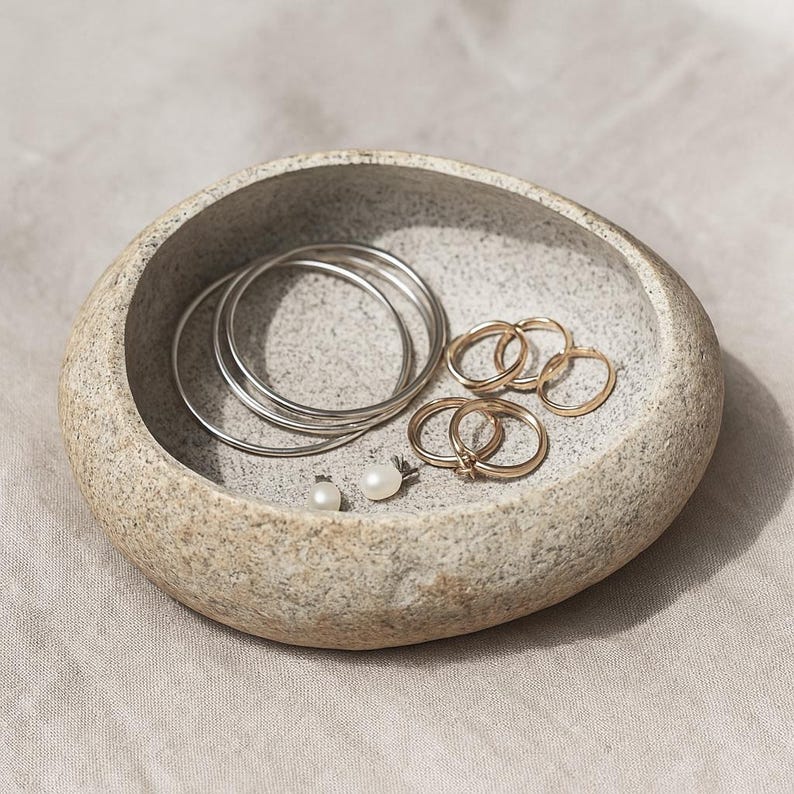 May include: A grey stone-effect jewellery dish contains silver bangles, gold rings, and pearl earrings. The dish has a textured surface and sits on a neutral fabric. The jewellery is arranged inside the dish.