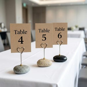 May include: Three table number holders with kraft paper cards. Each card reads "Table" and a number: 4, 5, and 6. The cards are held by heart-shaped wire clips attached to small, smooth stones. The stones are gray, tan, and black.