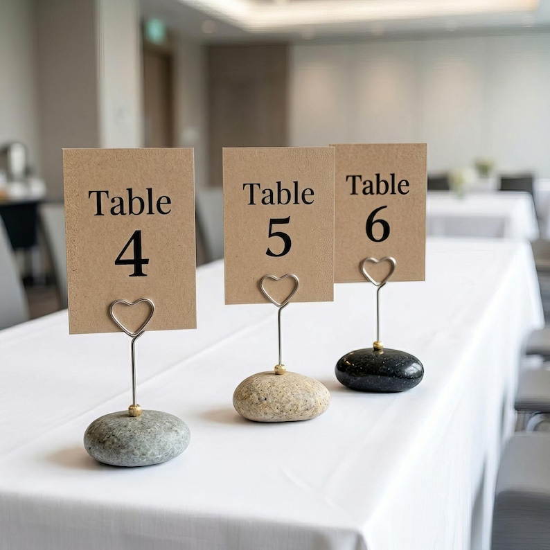 May include: Three table number holders with kraft paper cards. Each card reads "Table" and a number: 4, 5, and 6. The cards are held by heart-shaped wire clips attached to small, smooth stones. The stones are gray, tan, and black.